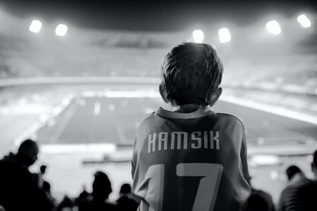 un enfant sur les épaules de son pere regarde un match de football et porte un maillot de foot rétro 