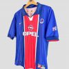 Photographie d’un maillot PSG 1998-1999 vintage emblématique. Le maillot est bleu avec une large bande rouge centrale, sponsorisé par Opel, avec le logo Nike et le blason du Paris Saint-Germain sur la poitrine, représentant un classique parmi les maillots de foot vintage de la fin des années 90.