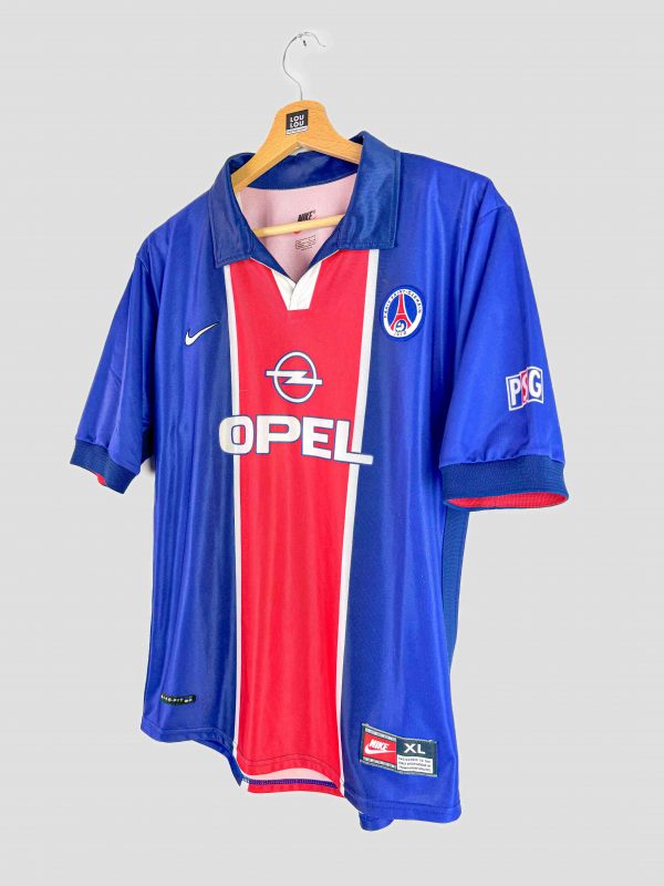 Photographie d’un maillot PSG 1998-1999 vintage emblématique. Le maillot est bleu avec une large bande rouge centrale, sponsorisé par Opel, avec le logo Nike et le blason du Paris Saint-Germain sur la poitrine, représentant un classique parmi les maillots de foot vintage de la fin des années 90.