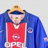 Vue de face d’un maillot du PSG de 1998, un modèle vintage signé Nike. La tunique de domicile présente un design bleu avec une bande rouge centrale emblématique, le sponsor Opel bien mis en avant sur le torse, et l’écusson du club parisien brodé sur la poitrine. Ce maillot de foot rétro incarne l’époque de la fin des années 90, avec ses couleurs et logos d’origine bien conservés.