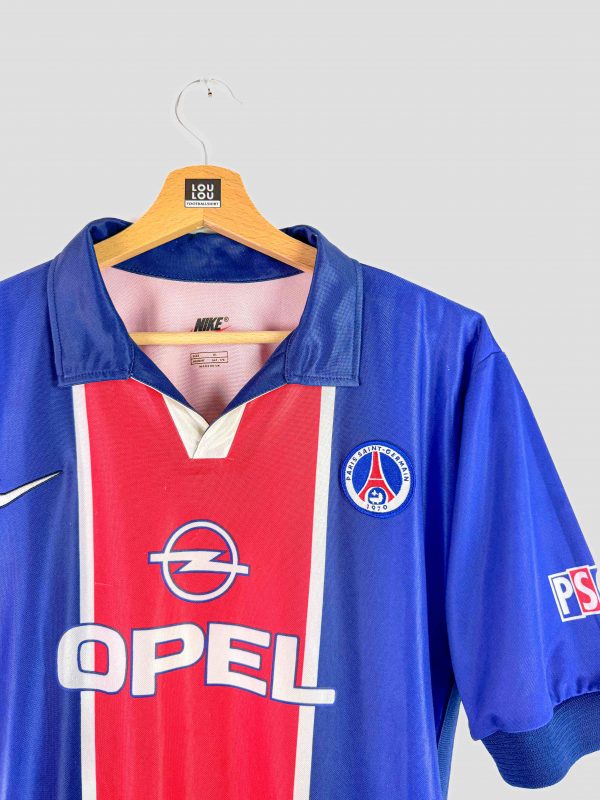 Vue de face d’un maillot du PSG de 1998, un modèle vintage signé Nike. La tunique de domicile présente un design bleu avec une bande rouge centrale emblématique, le sponsor Opel bien mis en avant sur le torse, et l’écusson du club parisien brodé sur la poitrine. Ce maillot de foot rétro incarne l’époque de la fin des années 90, avec ses couleurs et logos d’origine bien conservés.