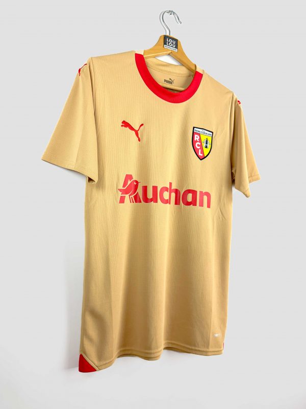 Le maillot away 2023-2024 du RC Lens, de couleur or avec le sponsor Auchan sur la poitrine et l’écusson du club en évidence.