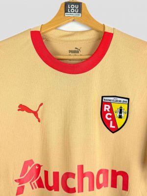 Nouveau maillot extérieur 2023/2024 du RC Lens, arborant un design doré unique avec logo Puma et inscription Auchan, présenté sur cintre.