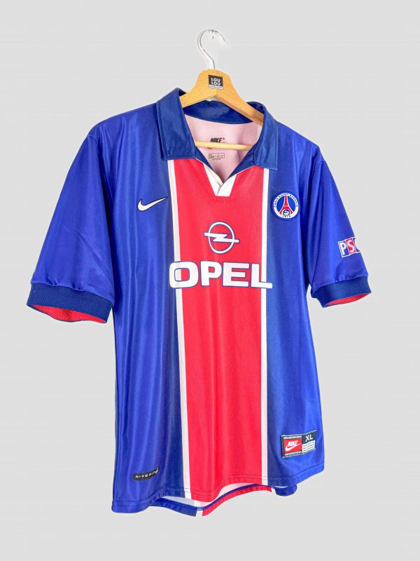 Image d’un maillot de football rétro du PSG datant de 1998. Le maillot domicile est bleu à bande rouge verticale, avec le logo Opel en sponsor central, le signe Nike sur le côté droit de la poitrine et le blason du PSG à gauche. Cette pièce classique des maillots de foot vintage est en très bon état.