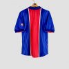 Photographie de dos d’un maillot du Paris Saint-Germain 1998-1999, pièce rétro emblématique du club. Le maillot est bleu avec sa bande rouge centrale caractéristique, il porte le sponsor Opel en blanc sur le torse, ainsi que le logo Nike et le blason du PSG. Cet ancien maillot de foot vintage symbolise l’époque 1998 du PSG et se présente dans un bel état de conservation.
