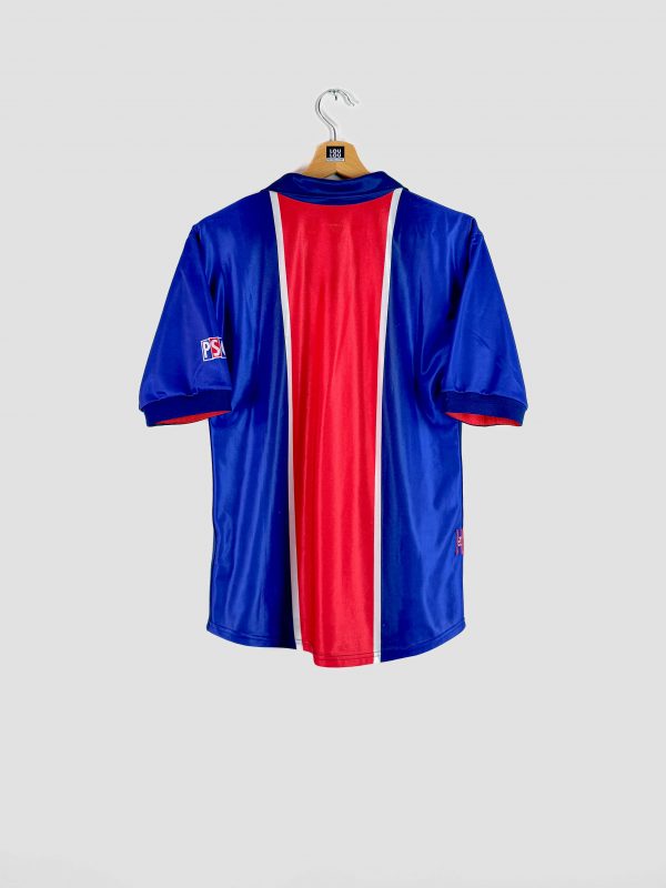 Photographie de dos d’un maillot du Paris Saint-Germain 1998-1999, pièce rétro emblématique du club. Le maillot est bleu avec sa bande rouge centrale caractéristique, il porte le sponsor Opel en blanc sur le torse, ainsi que le logo Nike et le blason du PSG. Cet ancien maillot de foot vintage symbolise l’époque 1998 du PSG et se présente dans un bel état de conservation.