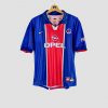 Photo d’un maillot de foot vintage du PSG (saison 1998-1999), de couleur bleue avec une large bande rouge centrale. Le maillot arbore le sponsor Opel en blanc sur la poitrine, le logo Nike et l’écusson du Paris Saint-Germain, et il est en excellent état de conservation.