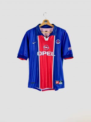 Photo d’un maillot de foot vintage du PSG (saison 1998-1999), de couleur bleue avec une large bande rouge centrale. Le maillot arbore le sponsor Opel en blanc sur la poitrine, le logo Nike et l’écusson du Paris Saint-Germain, et il est en excellent état de conservation.