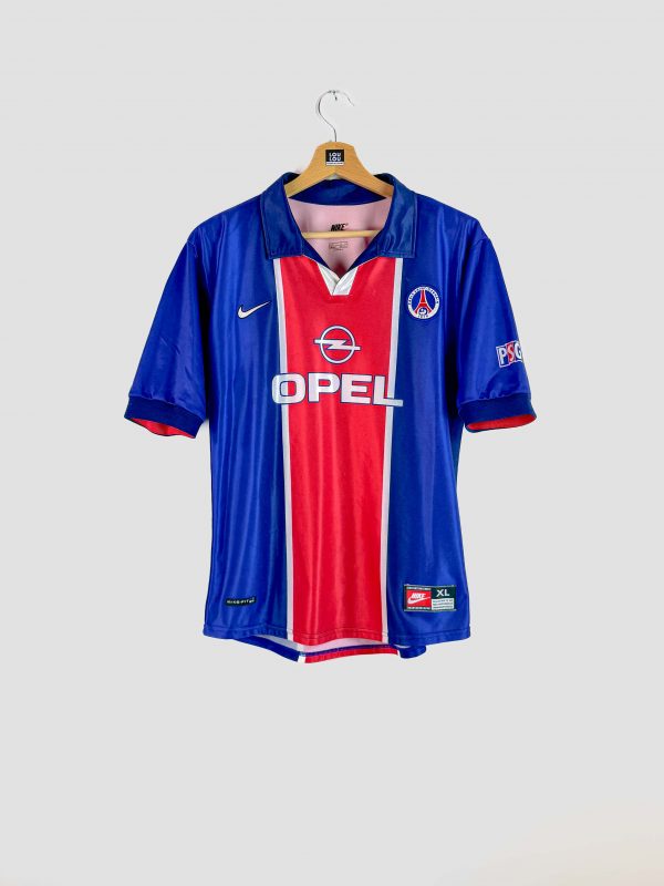 Photo d’un maillot de foot vintage du PSG (saison 1998-1999), de couleur bleue avec une large bande rouge centrale. Le maillot arbore le sponsor Opel en blanc sur la poitrine, le logo Nike et l’écusson du Paris Saint-Germain, et il est en excellent état de conservation.